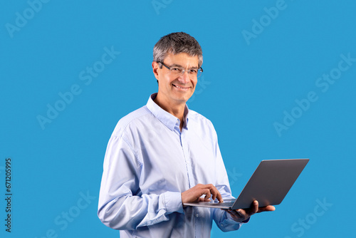 Smiling senior man browsing online on laptop in blue studio