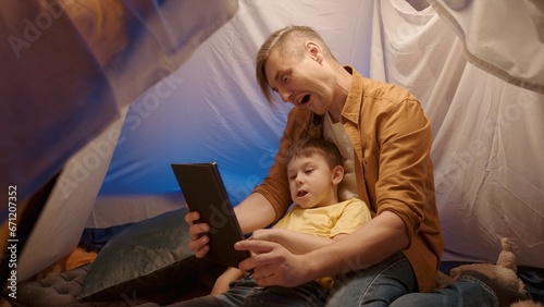 Father and son taking selfies with a tablet, making funny faces, close up. Father and son having fun in a camping tent decorated with garlands, in their living room in the evening.