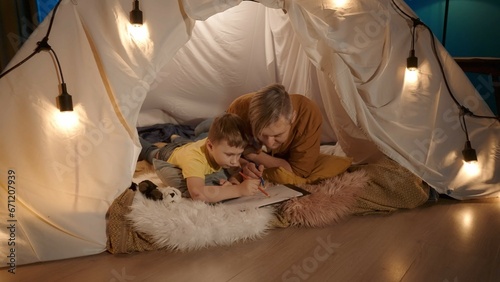 Dad and son are drawing together with colored pencils close up. Dad and son lying on their stomachs in a garland tent that is in the living room.