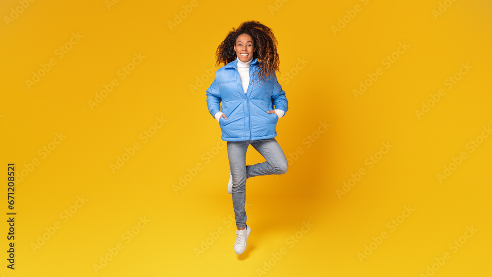 Young black lady wearing blue jacket, jumping against yellow backdrop