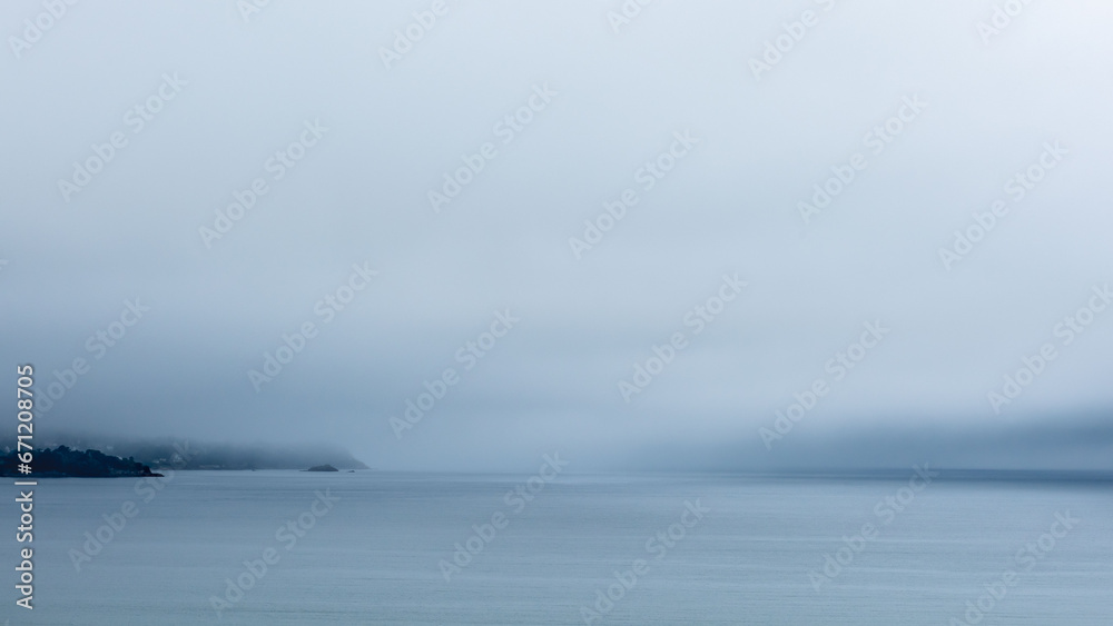 Une atmosphère mystérieuse en Bretagne avec une épaisse brume, des ciels gris, et la côte bretonne visible à l'horizon. La mer au premier plan est remarquablement calme, reflétant des tons gris et ble - obrazy, fototapety, plakaty 