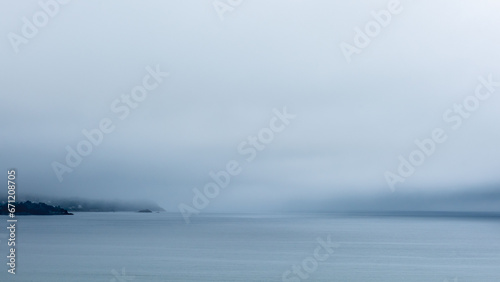 Une atmosphère mystérieuse en Bretagne avec une épaisse brume, des ciels gris, et la côte bretonne visible à l'horizon. La mer au premier plan est remarquablement calme, reflétant des tons gris et ble