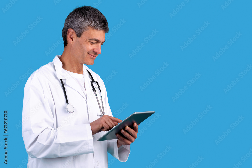 Senior physician websurfing on digital tablet posing over blue backdrop Senior physician websurfing on digital tablet posing over blue backdrop