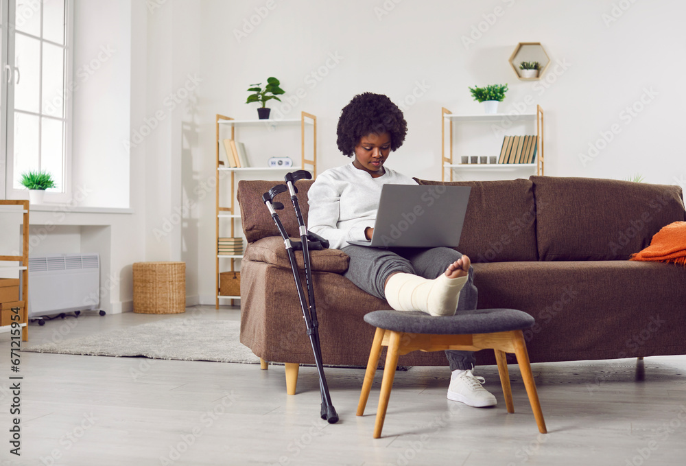 African American woman with fractured leg sitting on sofa and using laptop computer. Beautiful girl with plastered leg sitting on couch working online while recovering at home - obrazy, fototapety, plakaty 