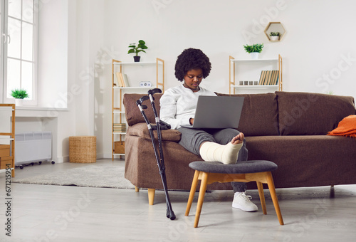 African American woman with fractured leg sitting on sofa and using laptop computer. Beautiful girl with plastered leg sitting on couch working online while recovering at home © Studio Romantic
