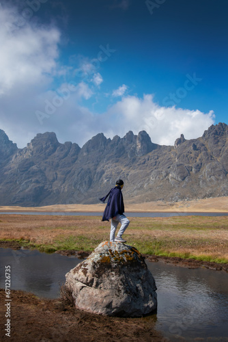 A lonely Peruvian man with black poncho and scarf standing on a large rock on the shores of a lake in the Andes mountains looking at the breadth of the landscape