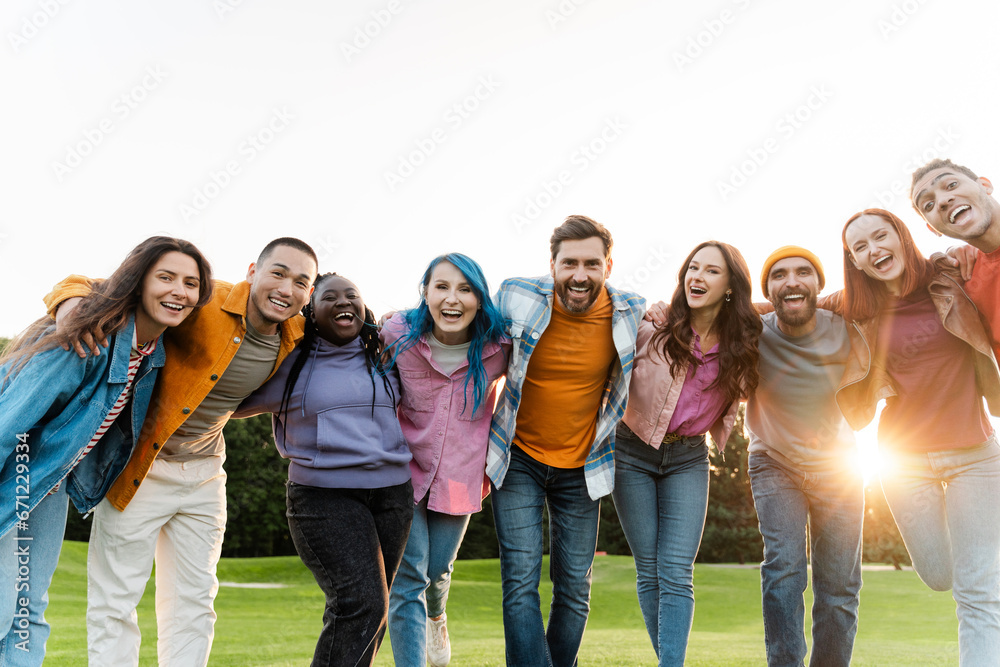 Group portrait of smiling multiracial friends hugging wearing colorful clothing looking at camera in park.  Happy attractive stylish students standing together outdoors . Diversity, friendship   