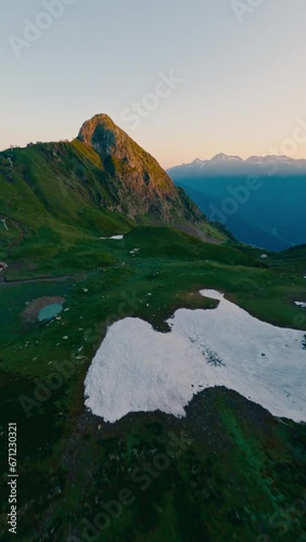 Vertical video 4k. Aerial view cinematic natural geology formation mountain valley green grass snow sunset. FPV sport drone rock alpine landscape sunny cliff summit peak altitude stone texture sunrise