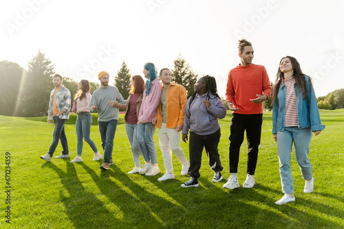 Group of smiling attractive multiracial friends wearing colorful clothing meeting, walking, talking in green park. Happy tourists together on vacation. Diversity, friendship, travel concept 
