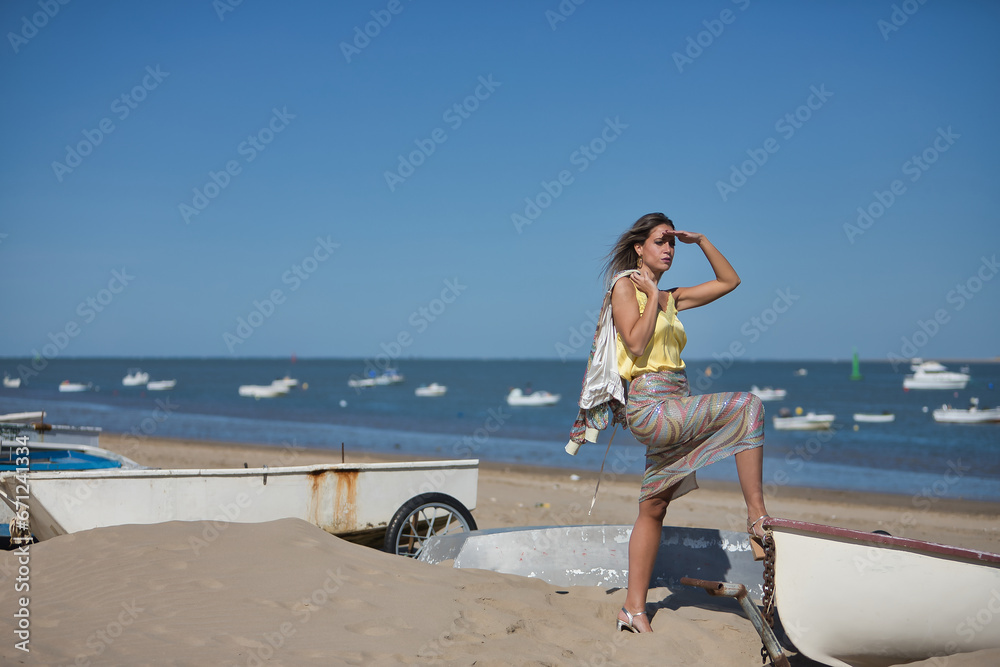 Young and beautiful woman in a sequined dress and high heels, with her foot on a boat on the beach, covering herself from the sun with her hand looking at infinity. Concept beauty, fashion, trend. Young and beautiful woman in a sequined dress and high heels, with her foot on a boat on the beach, covering herself from the sun with her hand looking at infinity. Concept beauty, fashion, trend.
