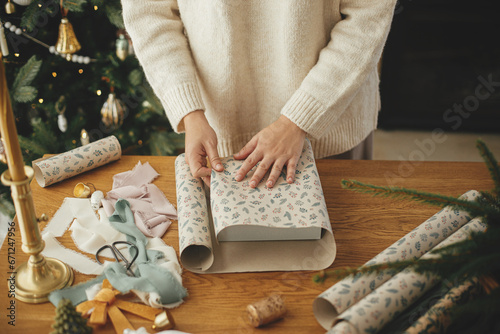 Woman in stylish sweater wrapping christmas gift in paper on wooden table with festive decorations in decorated scandinavian room. Merry Christmas! Hands packing present close up