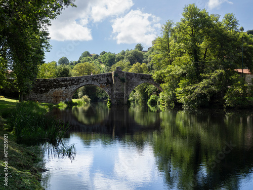 Puente antiguo de piedra con dos arcos sobre el río de Allaritz, reflejado en el agua junto al cielo y los árboles verdes alrededor de la orilla, viajando por Galicia en España, verano de 2021