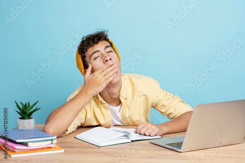 Attractive smiling teenager boy wearing casual clothes yawning sitting at desk using laptop photo
