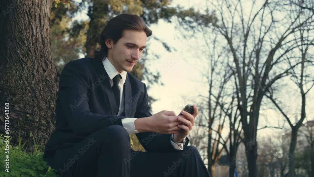 Smiling businessman in the city looking to a cell phone. Confident and happy man in a business suit is sitting outdoors and using smartphone. Young man in a suit with mobile phone. Smiling businessman in the city looking to a cell phone. Confident and happy man in a business suit is sitting outdoors and using smartphone. Young man in a suit with mobile phone.