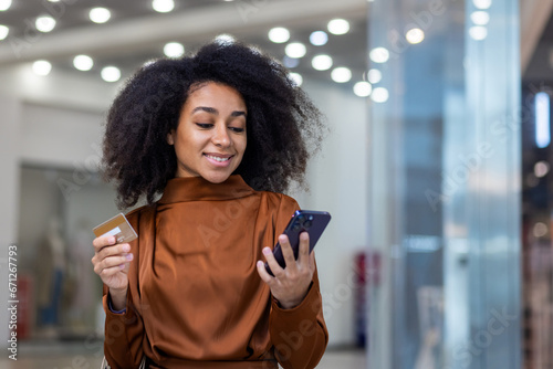 Young beautiful woman inside a supermarket large clothing store in a shopping mall using a smartphone app online shopping, an African American woman smiling contentedly holding a bank credit card. photo
