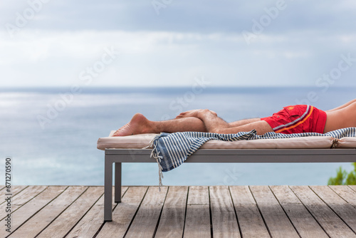 Obraz na plátně Les jambes d'un  homme en maillot de bain sur transat en terrasse, serviette pend du transat, mer et ciel  en arrière-plan