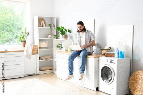 Young man with laptop sitting near washing machine at home
