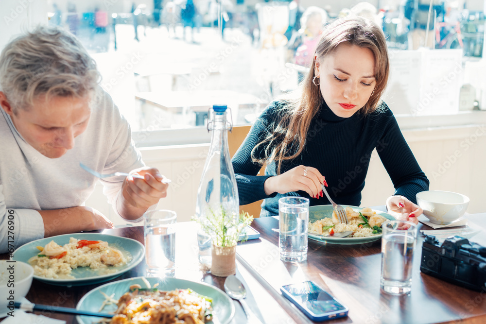 Young female and male friends sitting at served table in casual cafe and eating delicious hot risotto with shrimps or curry rice during lunch break. Trying new foods while traveling. real people. Young female and male friends sitting at served table in casual cafe and eating delicious hot risotto with shrimps or curry rice during lunch break. Trying new foods while traveling. real people.