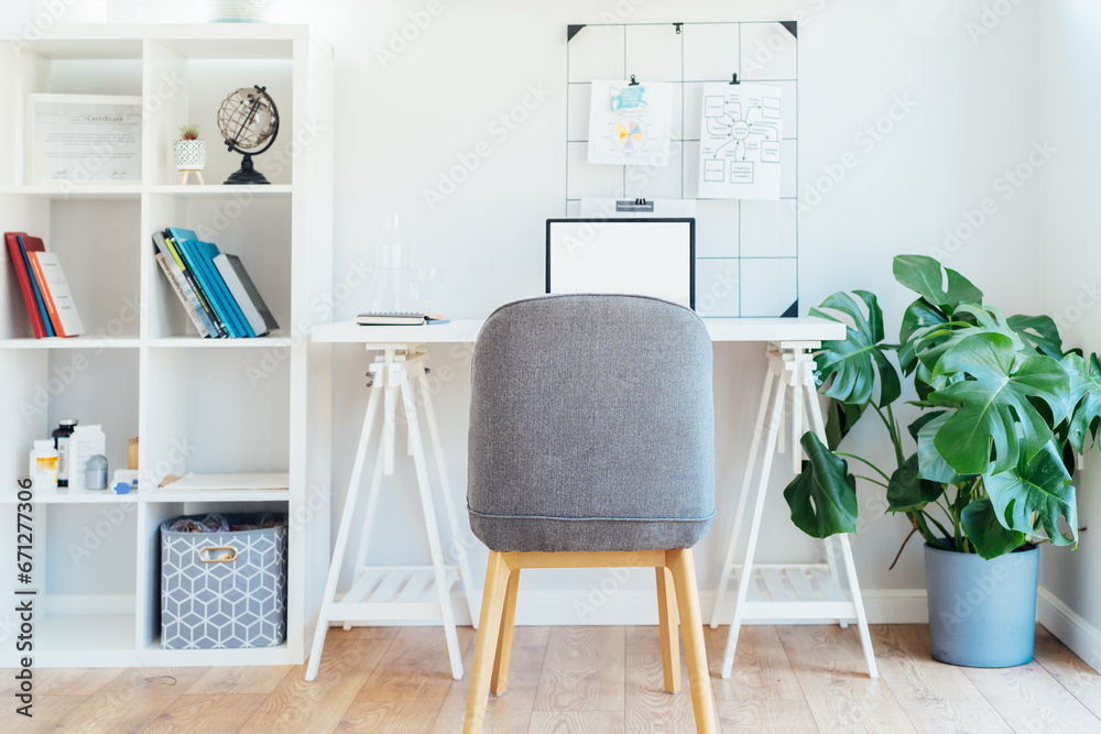 Fototapeta premium Modern cozy light workplace - white desk with laptop mockup empty screen, grid mood board with pinned notes, shelves with docs and green monstera plant at work space in home office room interior.