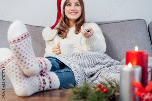 Hand holding remote controller with blurred smiling woman in santa hat, warm socks with cup of hot drink sitting on comfortable couch watching movie, TV with christmas decoration mood. Cozy winter