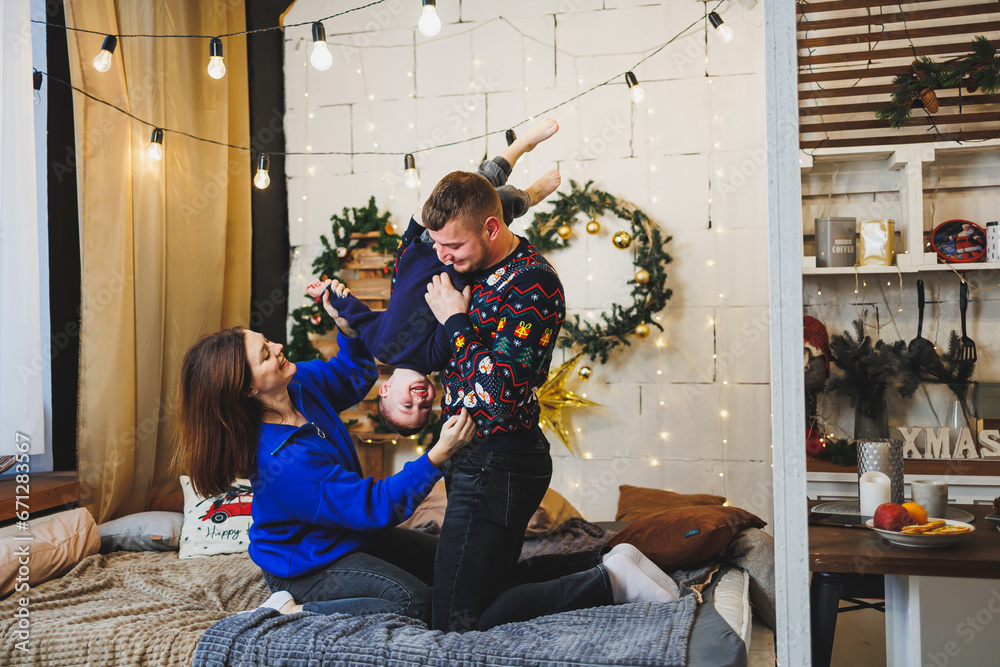 A cheerful young family with a small child rests near the Christmas tree at home. Family couple with Christmas decoration at home. Christmas, New Year - time to celebrate