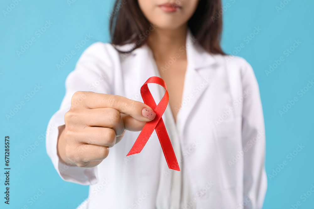 Young female doctor with red ribbon on blue background, closeup. World AIDS Day concept Young female doctor with red ribbon on blue background, closeup. World AIDS Day concept