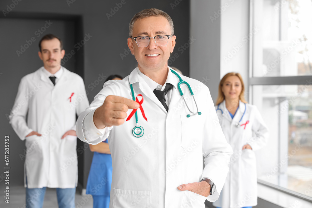 Mature male medic with red ribbon and group of doctors in clinic. World AIDS Day concept Mature male medic with red ribbon and group of doctors in clinic. World AIDS Day concept