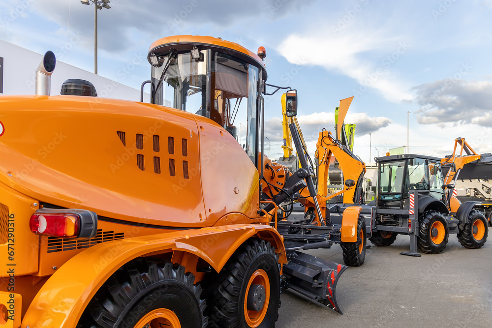 New orange wheel grader with rotary blade, rear view. Sale of new construction and municipal equipment at a dealership or fair. Heavy machinery