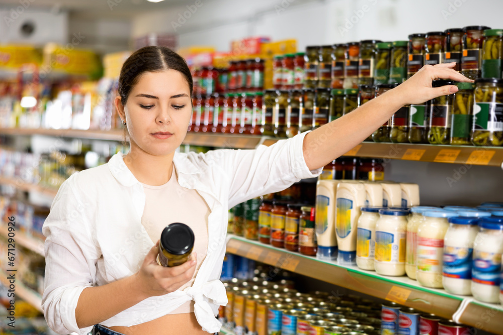 Focused interested young girl reading label on glass jar with canned goods while shopping in grocery section of supermarket