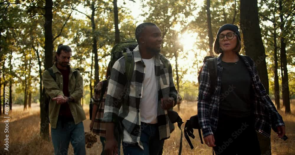 Happy international group on a hike in the summer forest. A man with Black skin in a checkered shirt and a girl with a bob with blond hair walk through the forest with their friends in special hiking