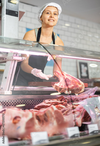 Sales girl in meat department of supermarket makes out showcase and puts pieces of meat on bones on shelf. Red meat, poultry, meat products and semi-finished products