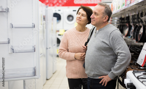 Mature man and woman standing in salesroom of appliance shop and choosing refrigerator.