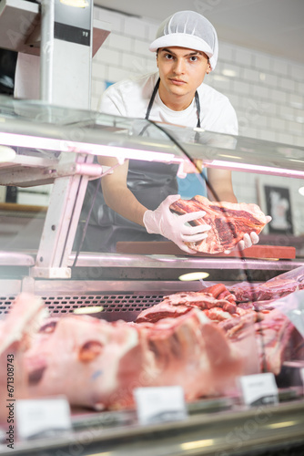 Young sales man holds piece of lamb loin and smiles at stores visitors. Guy employee takes meat on bones T-bone steak out of refrigerator window and prepares order for registration