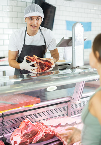 Positive young salesman standing behind counter demonstrating piece of meat to purchaser in butcher shop