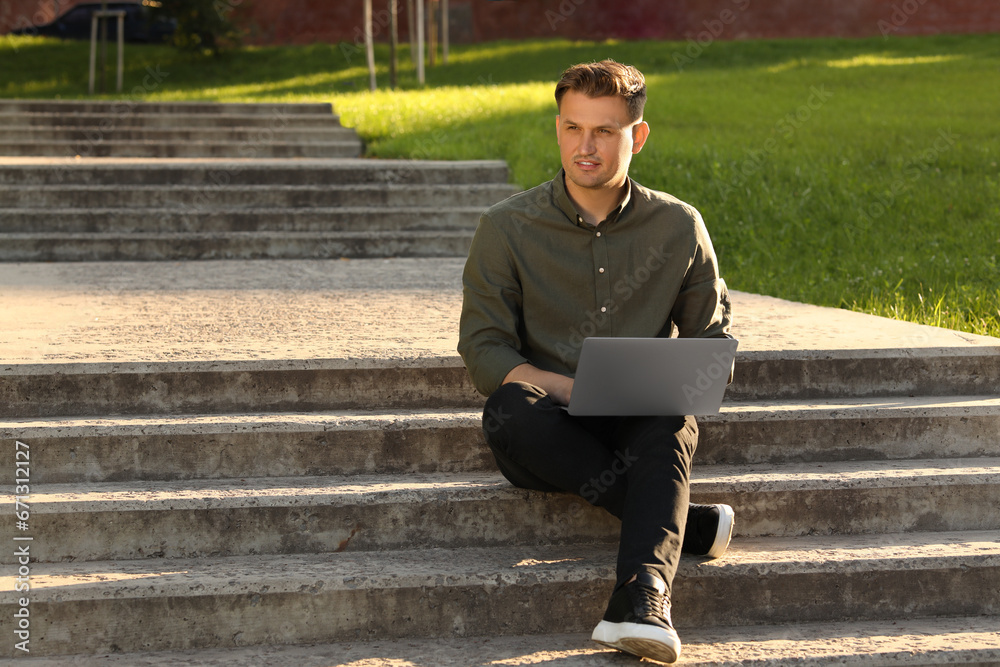 Obraz premium Handsome man with laptop sitting on concrete stairs outdoors. Space for text