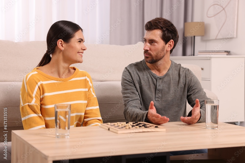 Happy couple talking while playing checkers at wooden table in room Happy couple talking while playing checkers at wooden table in room