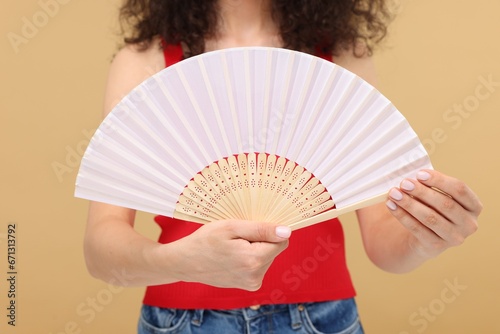 Woman holding hand fan on beige background, closeup