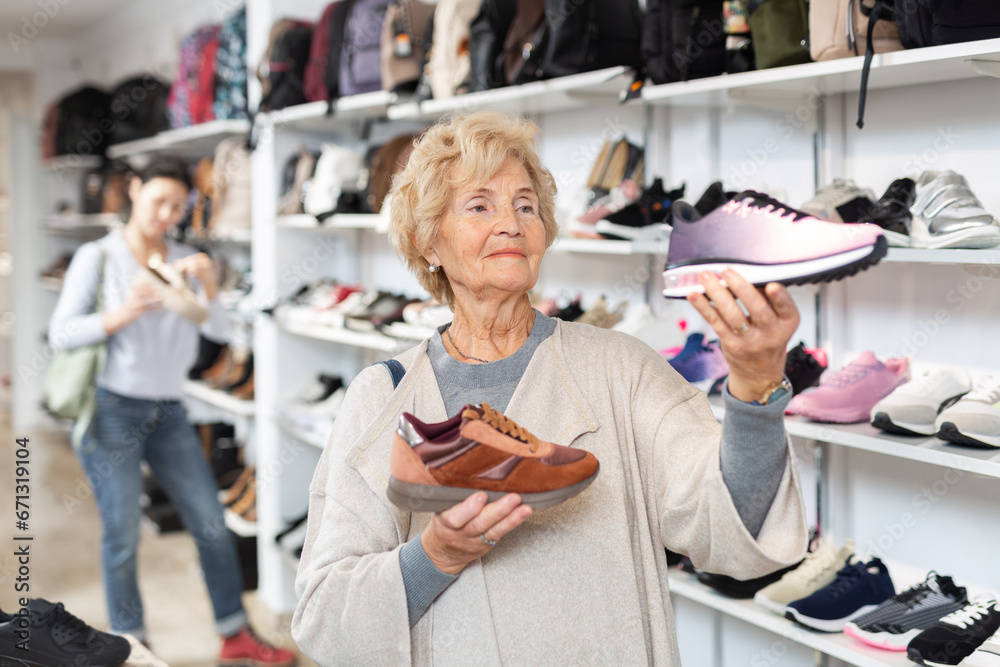 Mature european woman who came to a shoe store for shopping, chooses sports sneakers, standing near the shelves with the goods