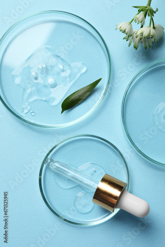 Petri dishes with samples of cosmetic oil, pipette and flowers on light blue background, flat lay