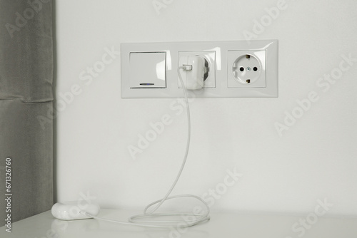 Charging case of modern wireless earphones plugged into power socket on white table indoors