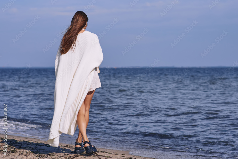Portrait of attractive brunette girl with long hair posing on a deserted beach. She wears white shorts, a top, a long white cape. She is looking to the camera. Portrait of attractive brunette girl with long hair posing on a deserted beach. She wears white shorts, a top, a long white cape. She is looking to the camera.