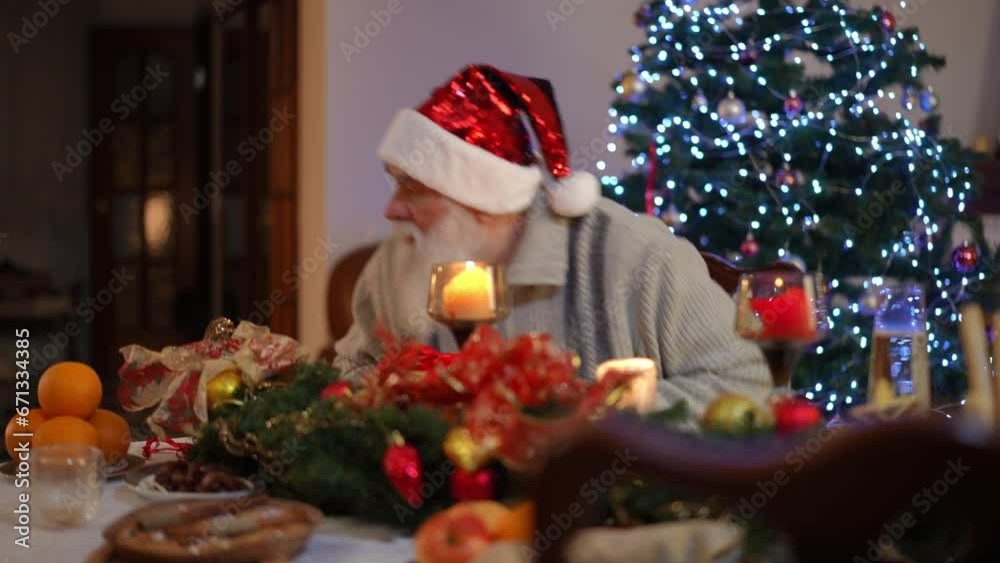 Sitting at the festive table near the Christmas tree, an elderly bearded man in a Christmas hat examines the gift box, and not finding a gift in it, gets up from the table and goes to look for it Sitting at the festive table near the Christmas tree, an elderly bearded man in a Christmas hat examines the gift box, and not finding a gift in it, gets up from the table and goes to look for it