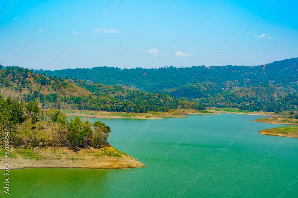 A serene man made lake surrounded by majestic hills, reflecting the blue sky and white clouds above in Tulungagung