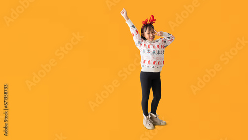 Cheerful young Asian girl wearing a Christmas sweater with reindeer horns, Happy smiling celebration success dancing and Hand up posing full body portrait, isolated on yellow background