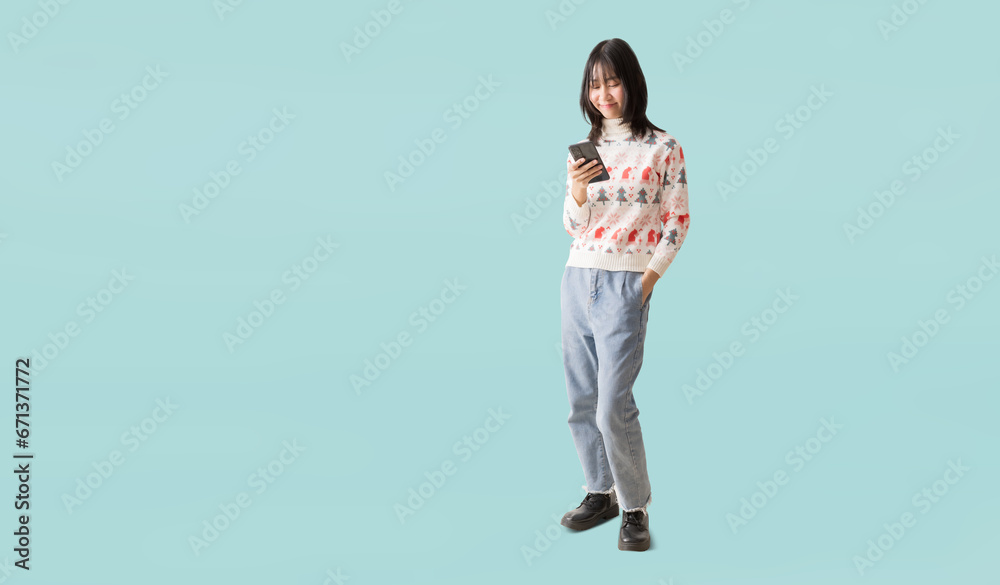 Happy smiling asian teenager girl standing using smart phone, Wearing a Christmas sweater, Portrait full body isolated on pastel plain light blue background