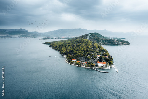 Aerial perspective of Marjan Hill in Split, Croatia. Verdant foliage dominates the peninsula, with anchored boats below and a moody sky above. A flock of birds graces the horizon.