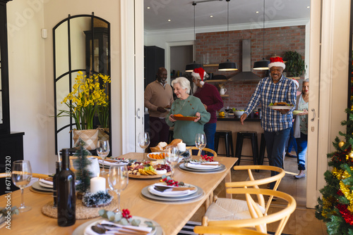 Happy diverse group of senior friends in santa hats serving food in sunny dining room at home