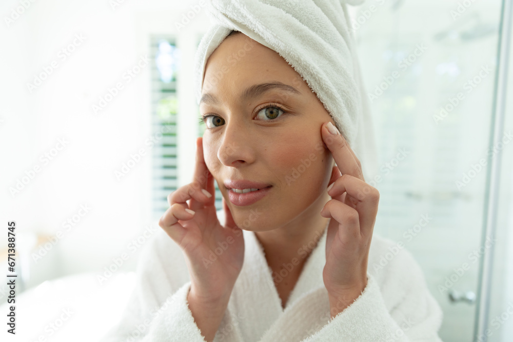 Happy caucasian woman in bathrobe looking in mirror, touching face and smiling in sunny bathroom Happy caucasian woman in bathrobe looking in mirror, touching face and smiling in sunny bathroom