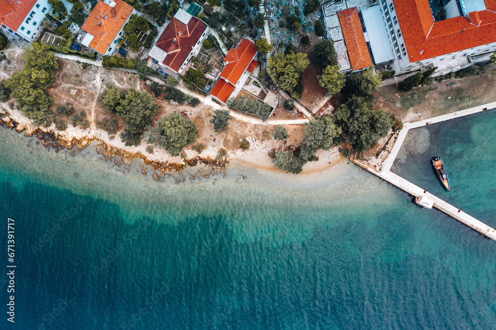 Aerial shot of Split's Marjan coastline, Croatia: Coastal homes with terra-cotta roofs flank a rustic path, leading to a pier with a moored boat against a gradient of blue waters.