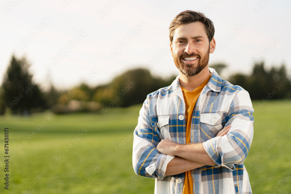 Portrait of smiling handsome middle aged man farmer wearing s stylish t shirt holding arms crossed looking at camera standing in green field, copy space. Successful business concept - obrazy, fototapety, plakaty 